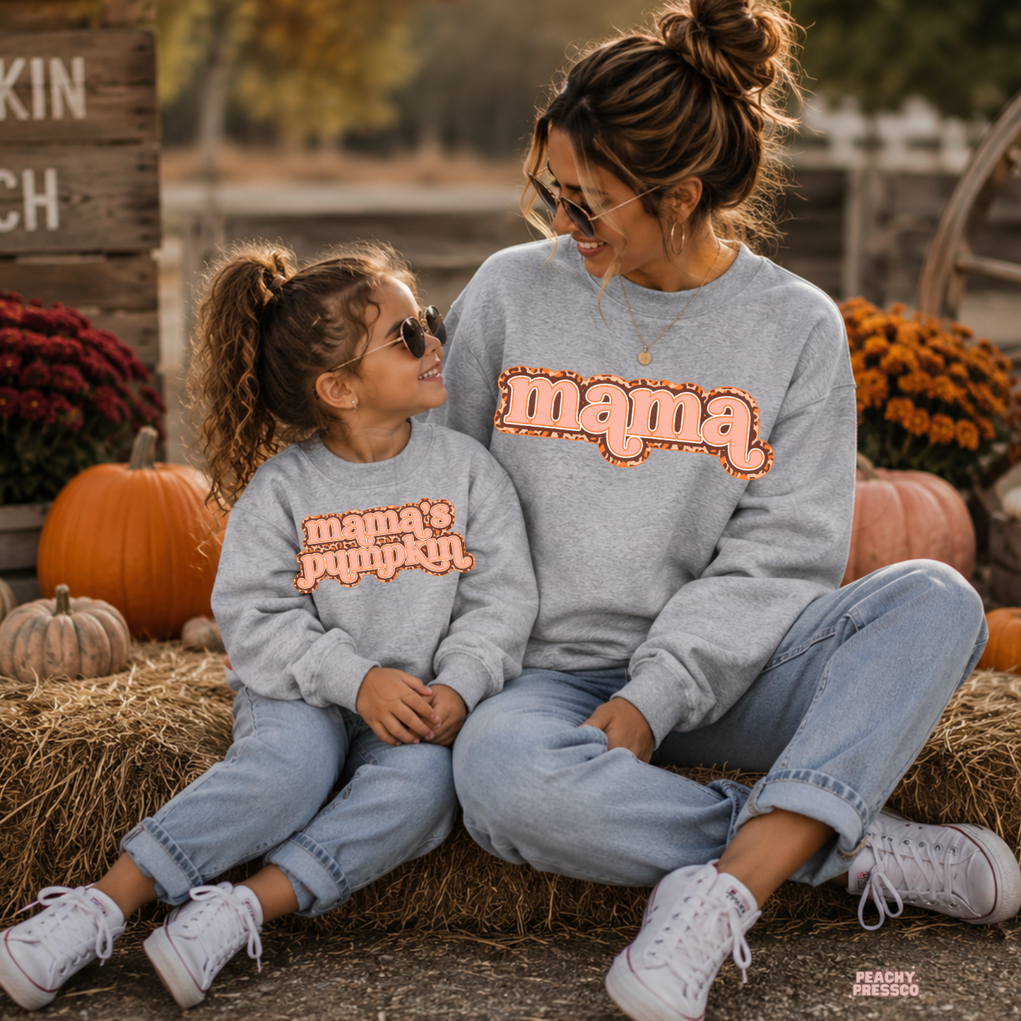 Mama and daughter wearing matching sport grey sweatshirts with "Mama" and "Mama's Pumpkin" designs while sitting together at a pumpkin patch.