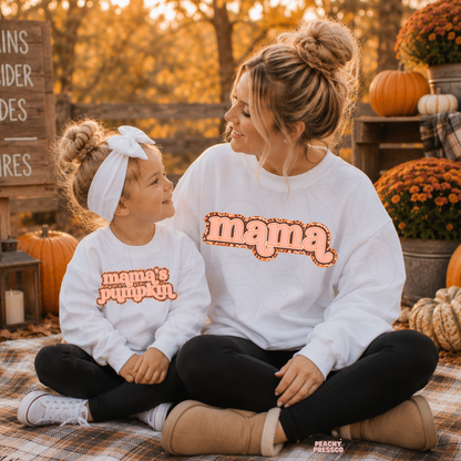 Mother and daughter smiling at each other wearing matching white "Mama" and "Mama's Pumpkin" fall sweatshirts with cozy autumn decor.