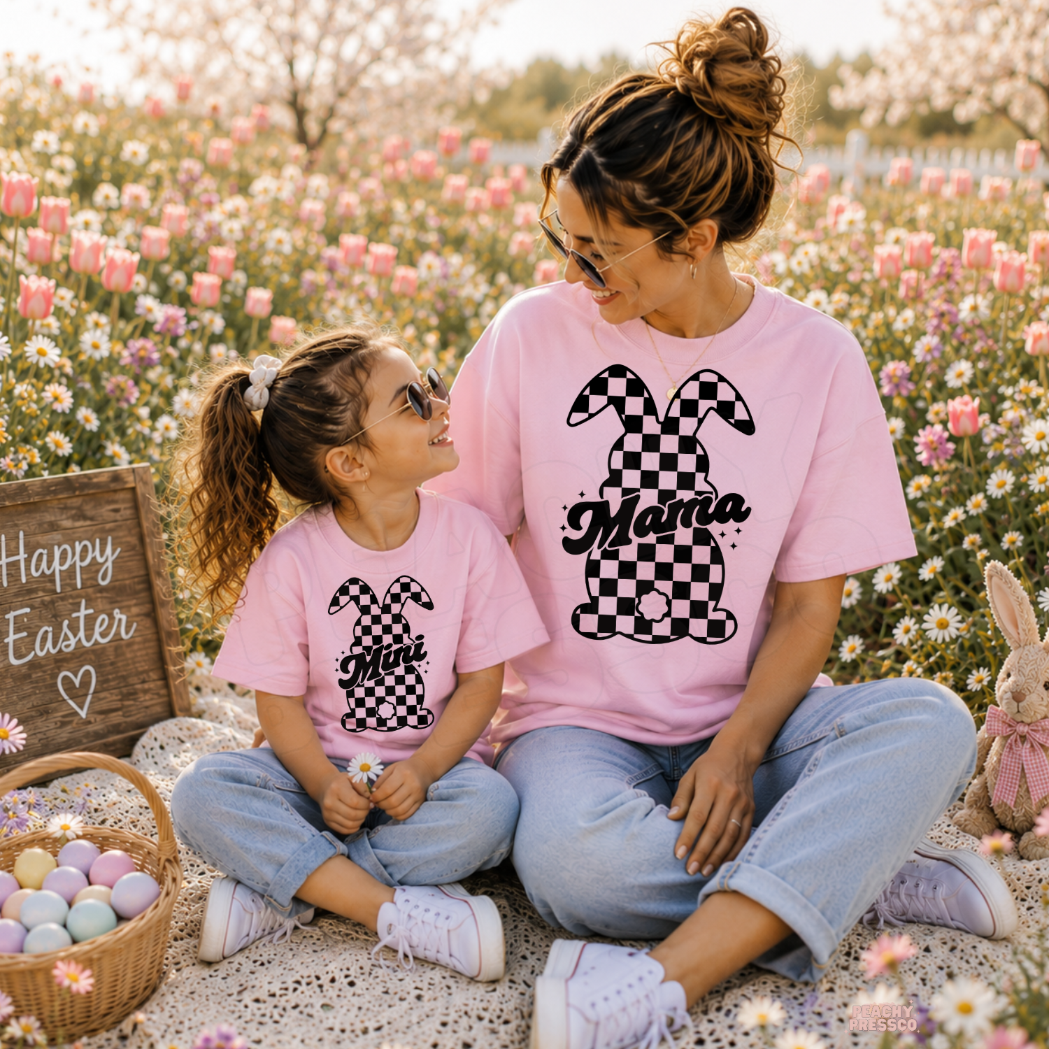 Mom and daughter wearing matching light pink checkered bunny Mama and Mini Easter shirts in a spring flower field with Easter eggs.