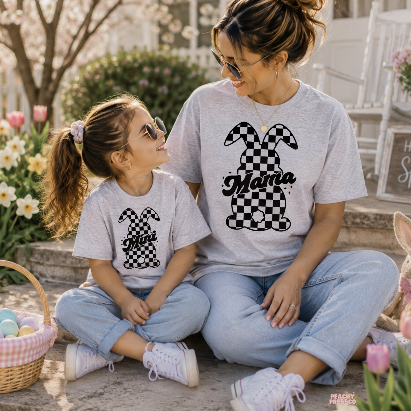 Mother and daughter wearing matching ash grey checkered bunny Mama and Mini Easter t-shirts while sitting outside in a spring garden.