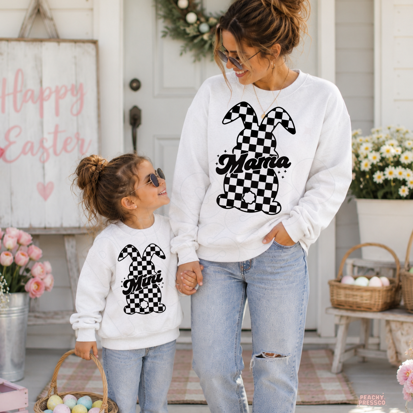 Matching white Mama and Mini checkered bunny Easter crewneck sweatshirts for mom and daughter holding hands outside a spring farmhouse.