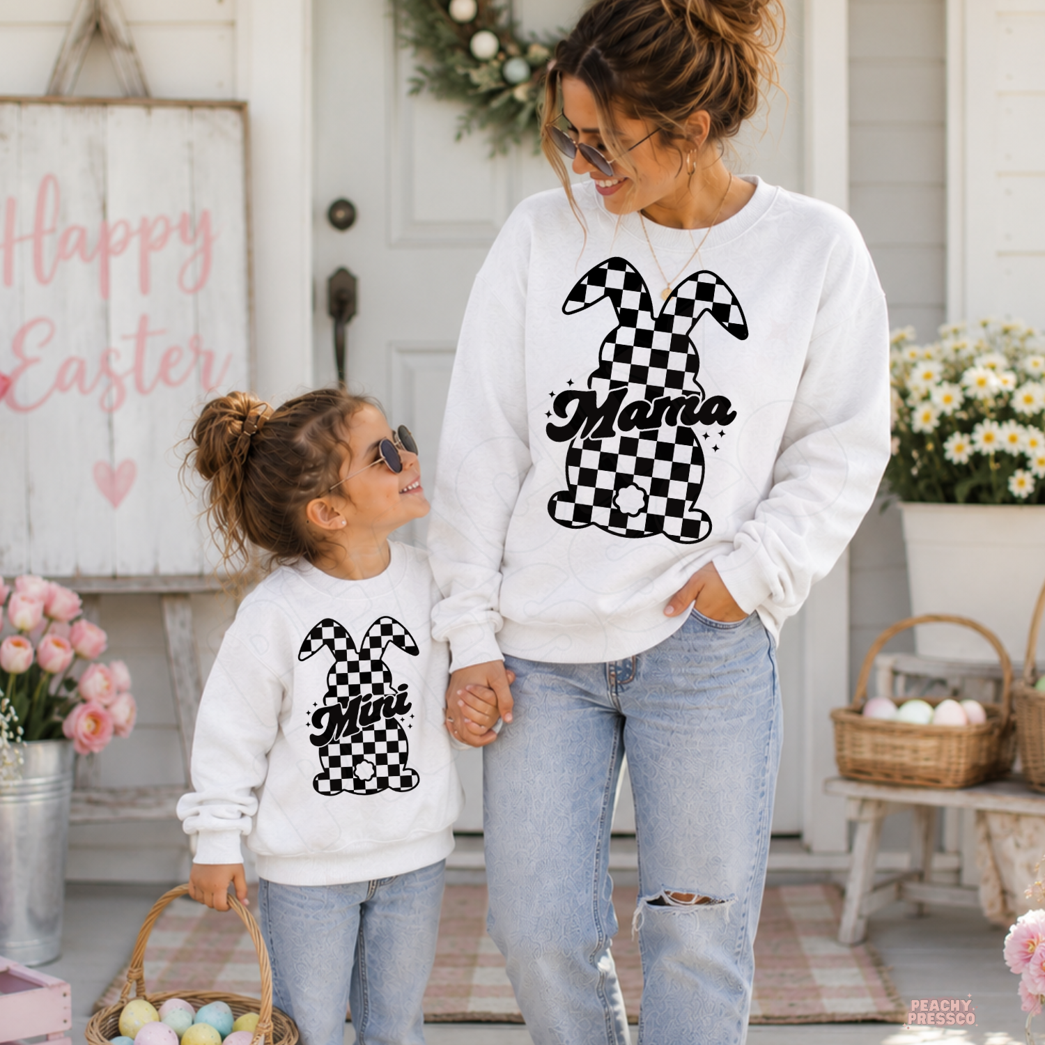 Matching white Mama and Mini checkered bunny Easter crewneck sweatshirts for mom and daughter holding hands outside a spring farmhouse.