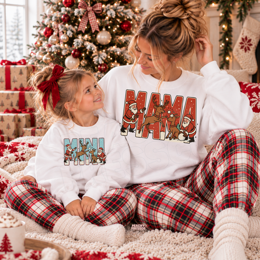 Mom and daughter wearing matching white Mama and Mini Christmas sweatshirts featuring a retro Santa and reindeer design.