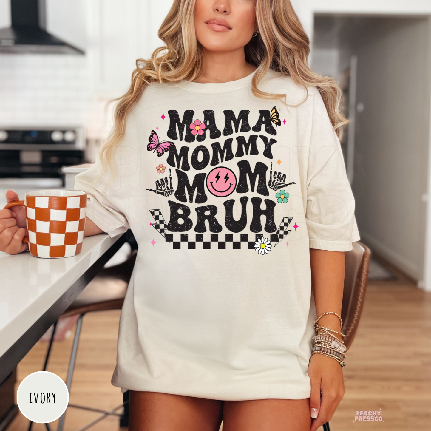 Ivory graphic t-shirt featuring a fun "Mama Mommy Mom Bruh" retro design with distressed lettering, colorful flowers, butterflies, and checkerboard pattern.