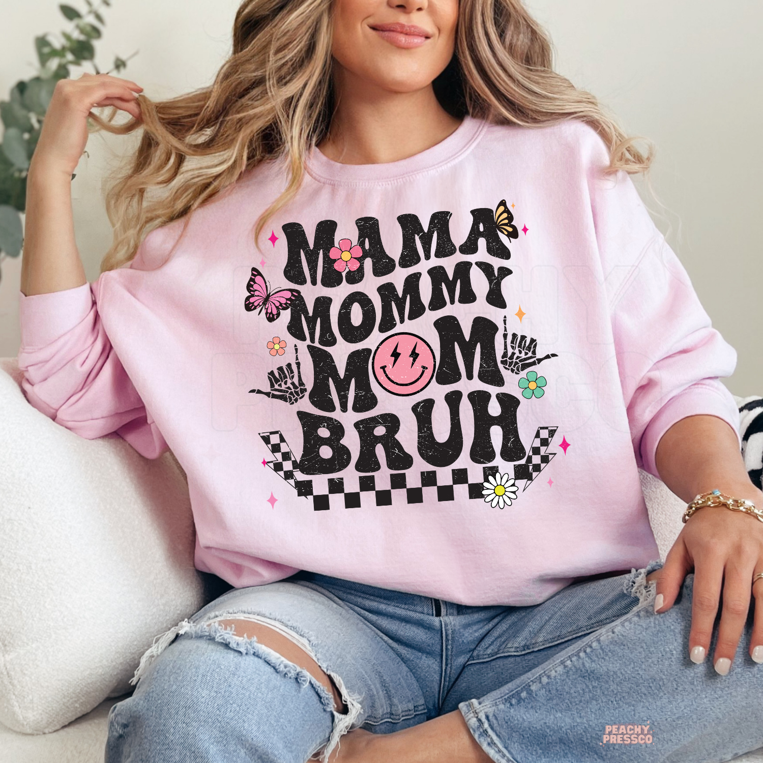 Light pink sweatshirt featuring a trendy "Mama Mommy Mom Bruh" graphic with retro typography, smiley face, floral accents, and butterflies. Cute mom gift.