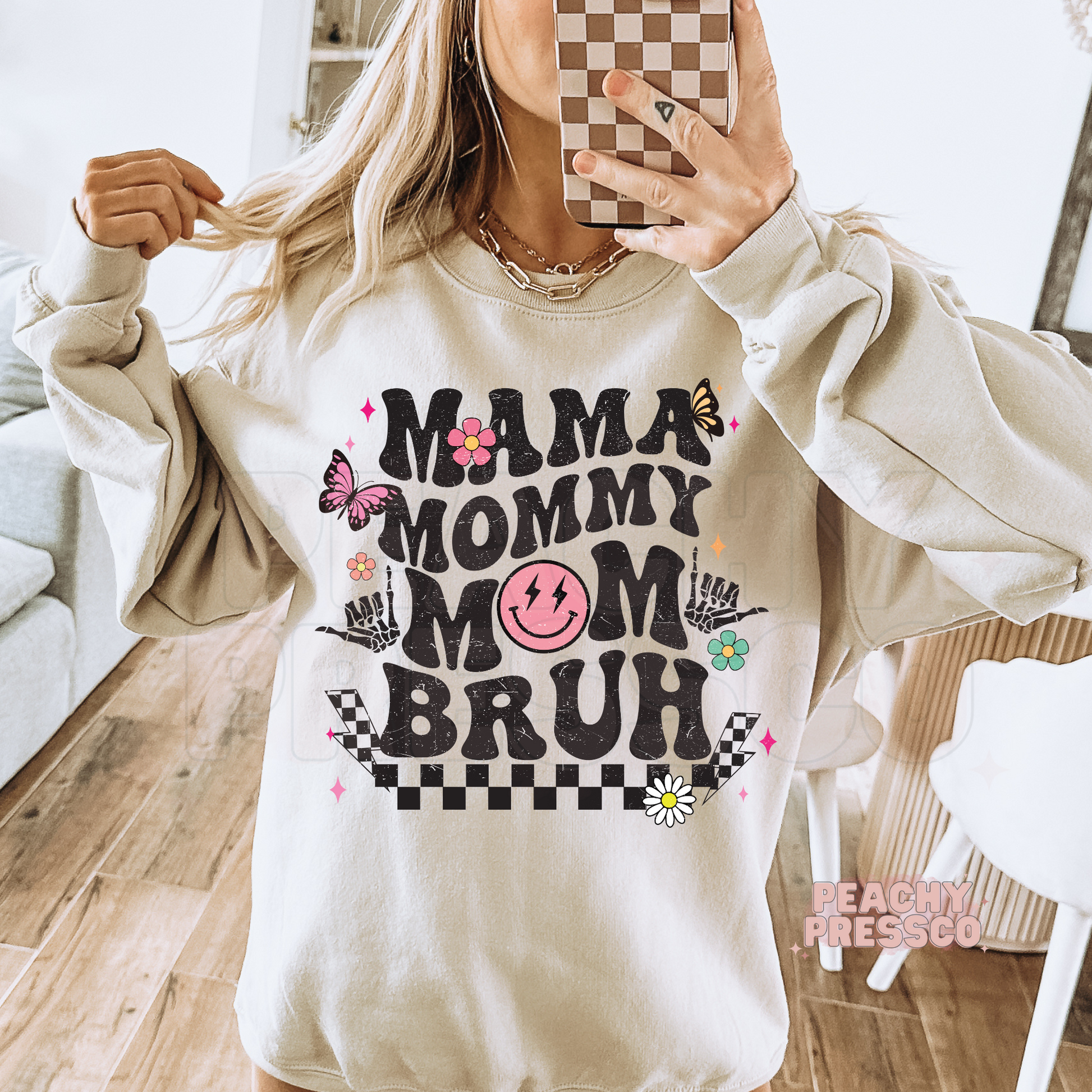 Sand sweatshirt featuring a retro "Mama Mommy Mom Bruh" design with distressed lettering, checkerboard pattern, flowers, and butterflies.