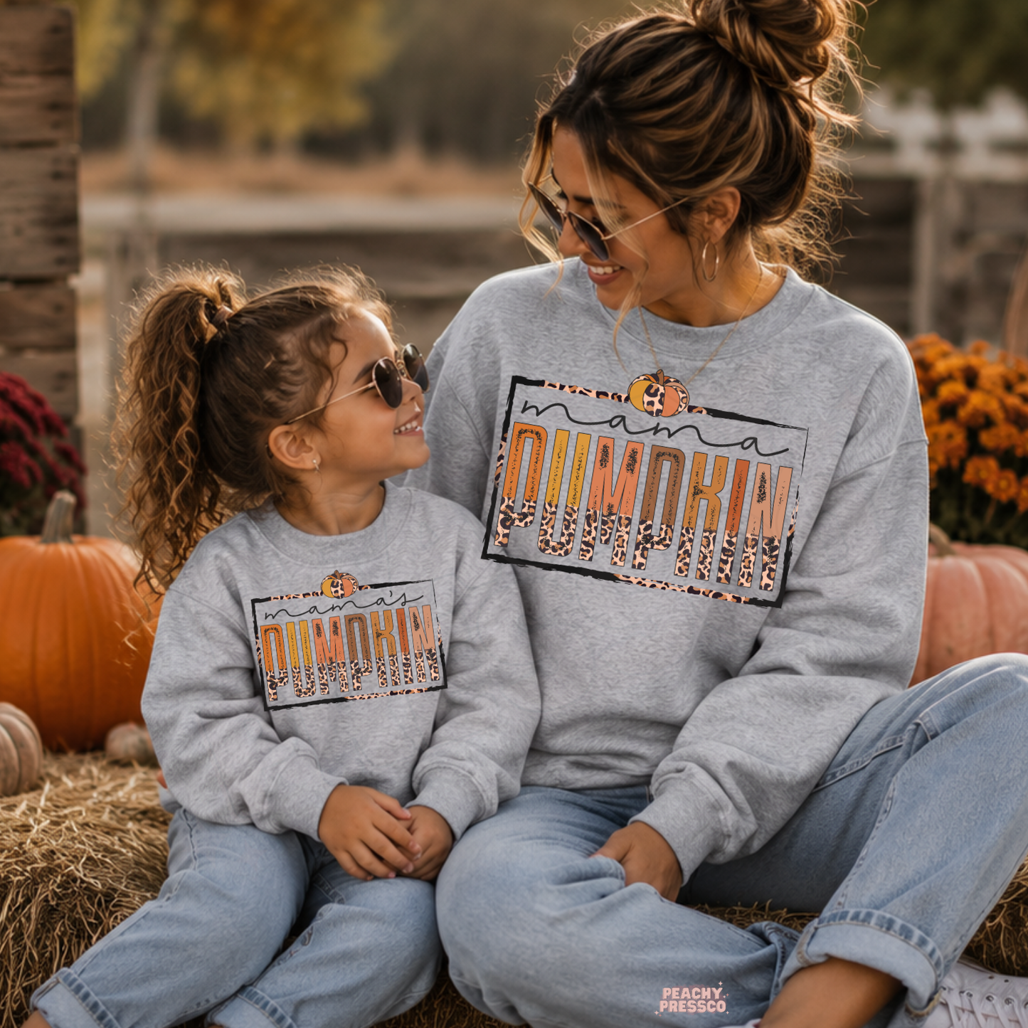 Mother and daughter in matching sport grey Mama Pumpkin sweatshirts sitting together at a pumpkin patch with pumpkins and fall decorations.
