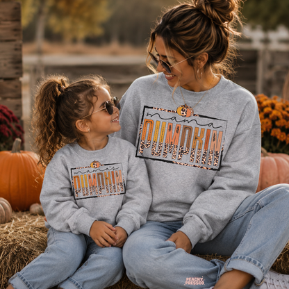 Mother and daughter in matching sport grey Mama Pumpkin sweatshirts sitting together at a pumpkin patch with pumpkins and fall decorations.