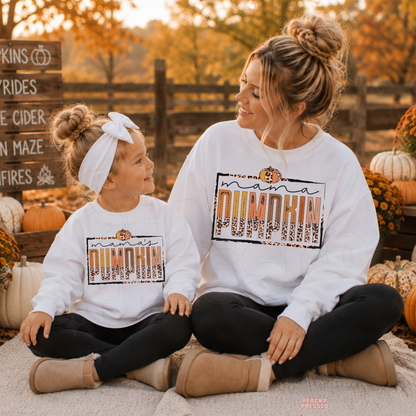 Mom and daughter wearing matching white Mama Pumpkin sweatshirts at a pumpkin patch, smiling together during a cozy autumn day.