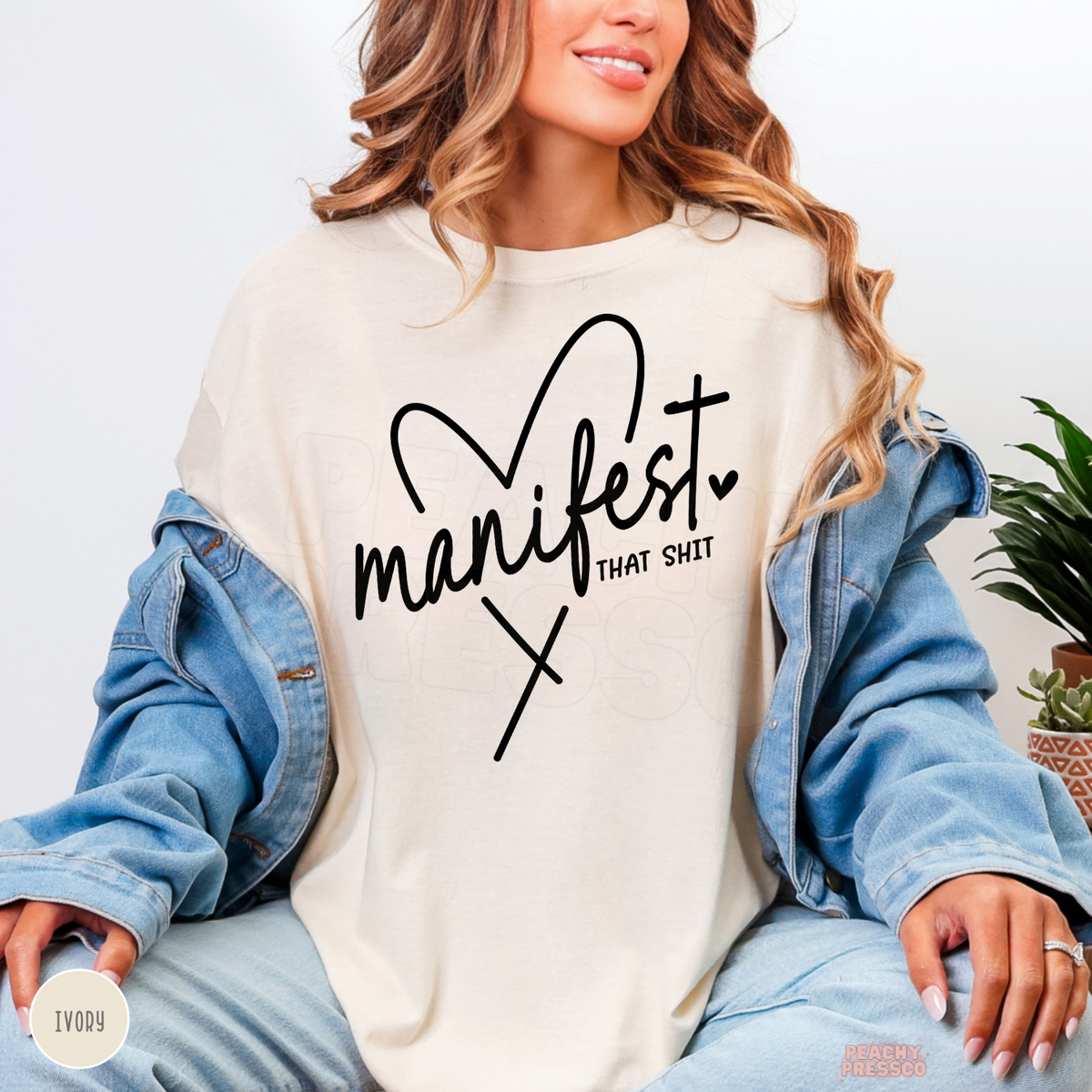 Ivory Manifest That Shit shirt featuring black handwritten typography, a heart accent and a sassy inspirational quote about manifesting dreams.