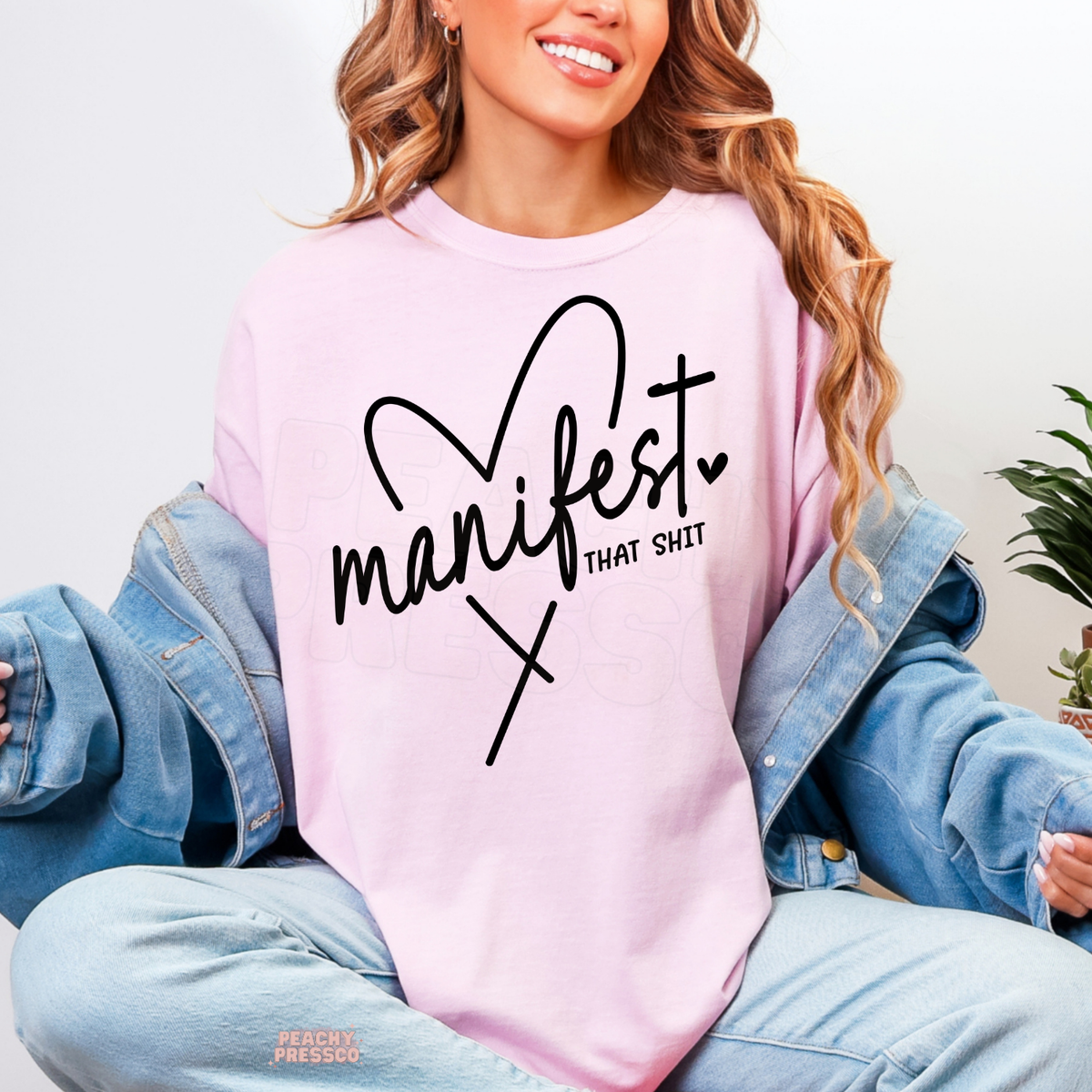 Light pink Manifest That Shit shirt featuring black handwritten script, a cute heart detail and a funny positive mindset quote.