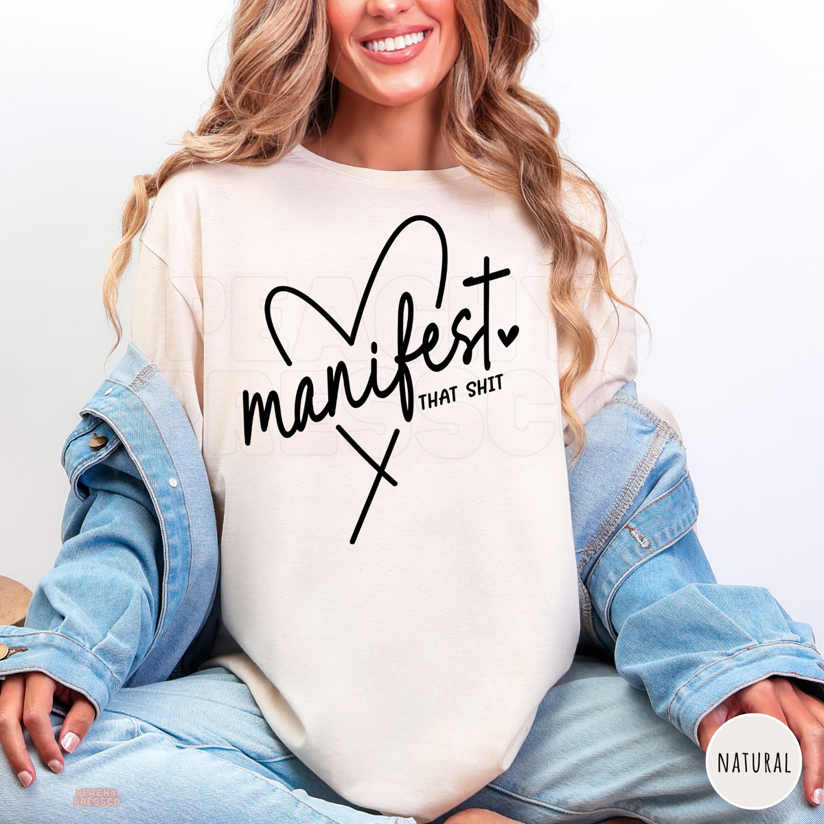 Natural Manifest That Shit shirt featuring black handwritten lettering, a heart accent and a bold motivational manifestation quote.