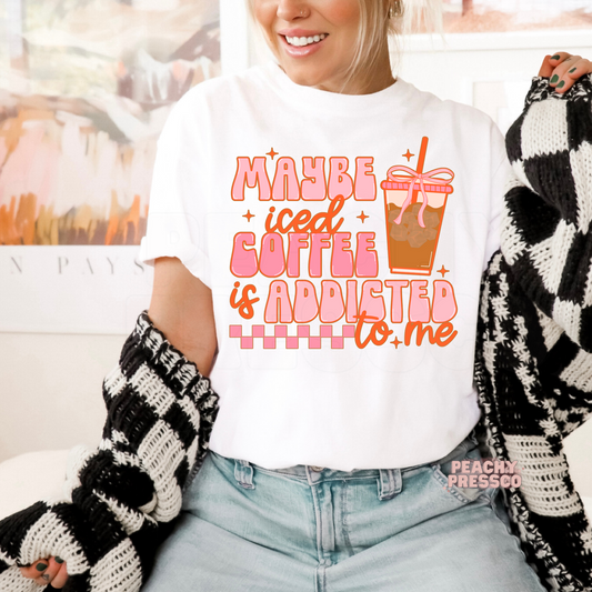 Woman wearing a white graphic t-shirt featuring the funny "Maybe Iced Coffee Is Addicted to Me" design with a pink iced coffee cup illustration.