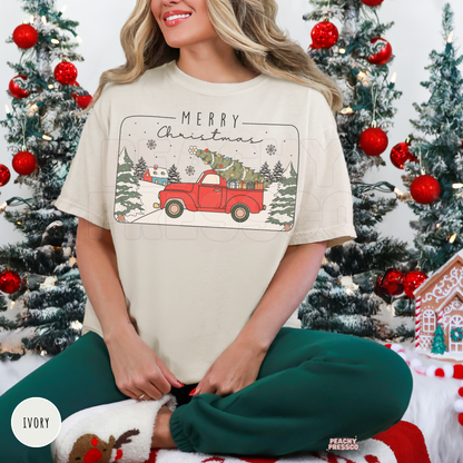 Woman sitting in front of Christmas trees wearing an ivory t-shirt featuring a vintage red pickup truck carrying a Christmas tree.