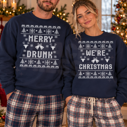 A man and a woman wearing matching navy blue crewneck sweatshirts with white holiday-themed graphics. The man's sweatshirt reads 'MERRY DRUNK' and the woman's reads 'WE'RE CHRISTMAS', both decorated with white snowflakes, Christmas trees, and gift boxes.