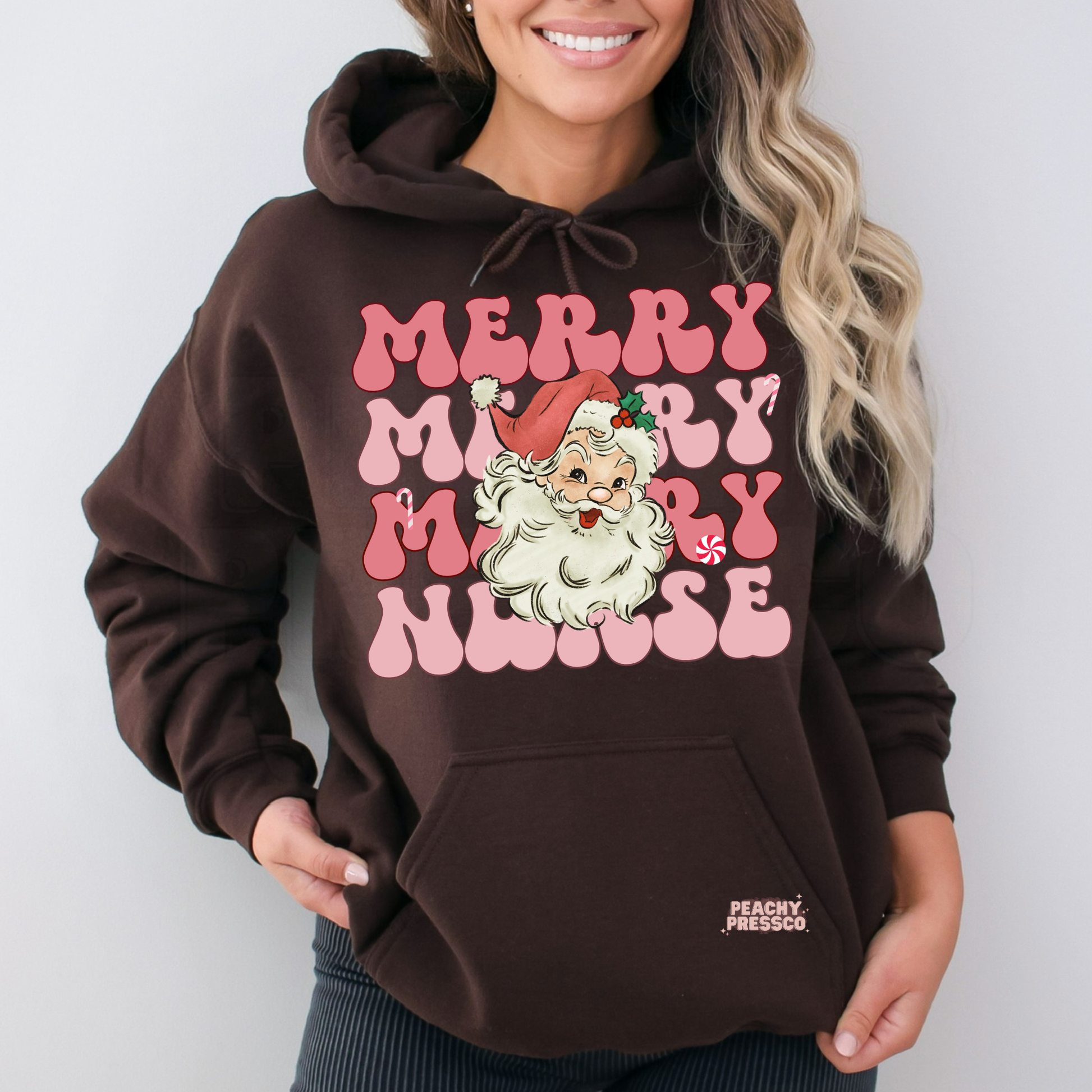 A woman wearing a dark brown pullover hoodie with a 'Merry Merry Nurse' graphic featuring a cartoon Santa Claus and pink lettering, on a light gray background.
