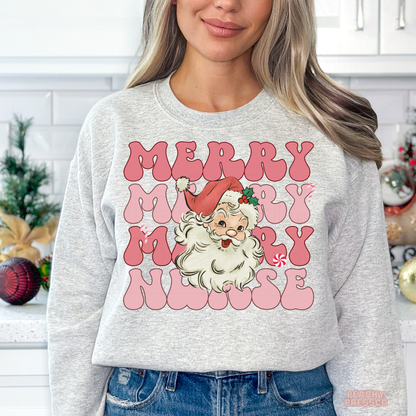 A woman wearing a ash grey sweatshirt with a festive graphic that reads 'MERRY MERRY NURSE' in pink bubble letters, featuring a cartoon Santa Claus in the center. The background shows a softly blurred kitchen setting with holiday decorations.