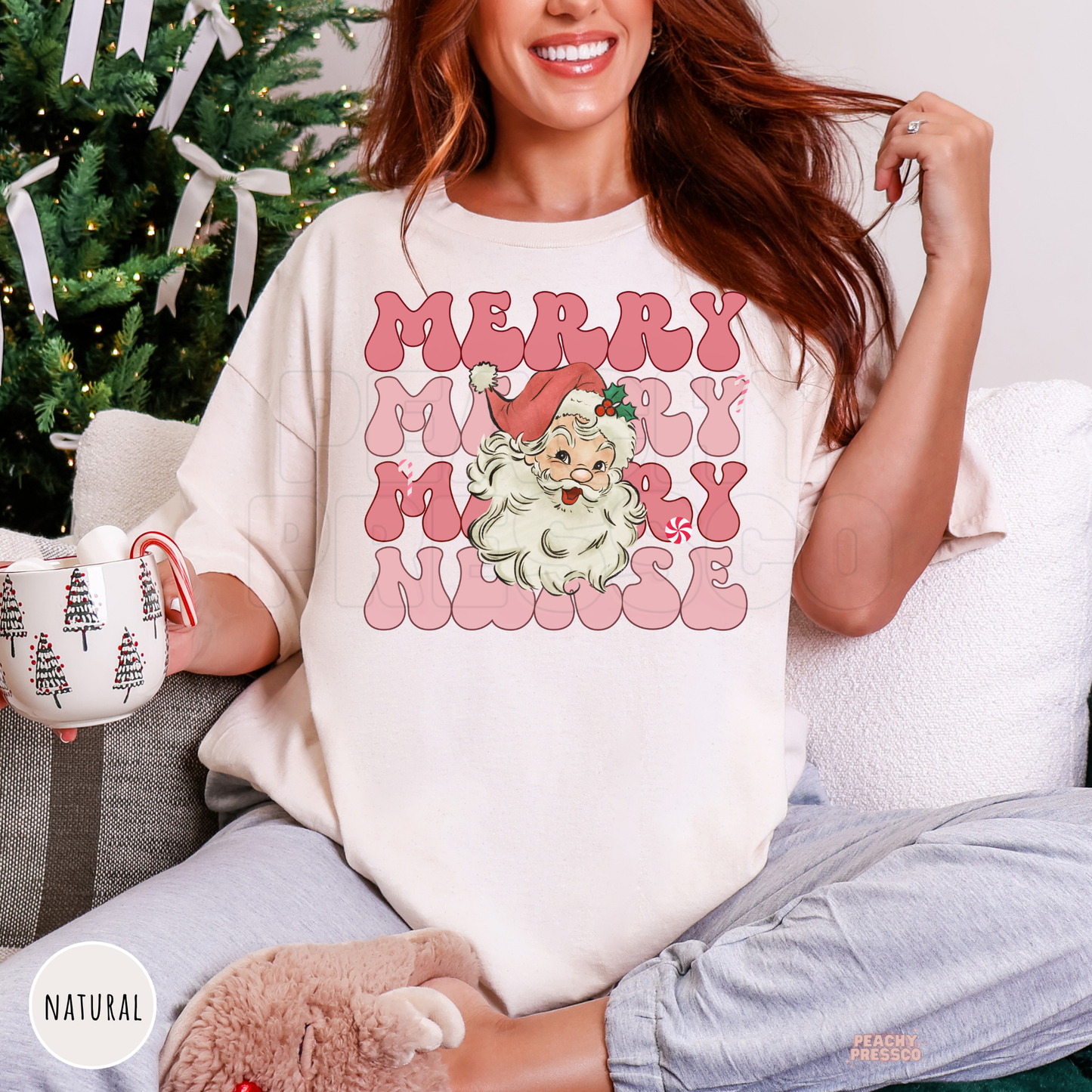 A woman sitting on a couch wearing an oversized natural colored t-shirt with a pink and white graphic that reads 'MERRY MERRY NURSE' with a vintage Santa. She is holding a mug in front of a decorated Christmas tree.