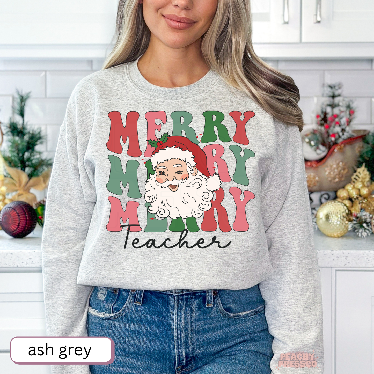 A woman wearing an ash grey crewneck sweatshirt with a festive 'Merry Merry Merry Teacher' graphic featuring a Santa Claus face. The background shows a decorated holiday kitchen with Christmas ornaments and a sleigh.
