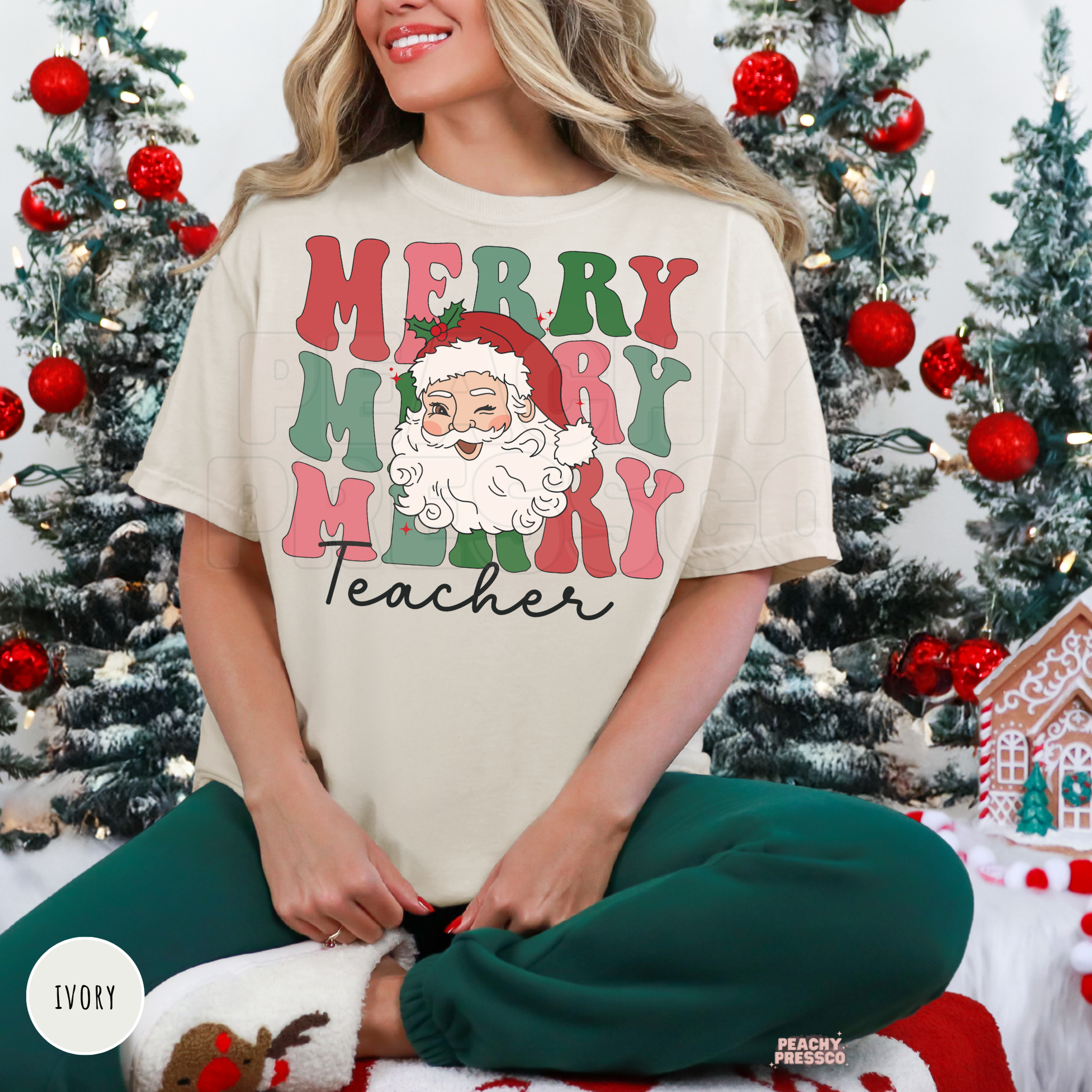A woman wearing an ivory graphic t-shirt with a Christmas 'Merry Teacher' design, sitting in front of decorated Christmas trees. The t-shirt features a colorful retro-style graphic with Santa Claus and the text 'MERRY MERRY MERRY Teacher'.