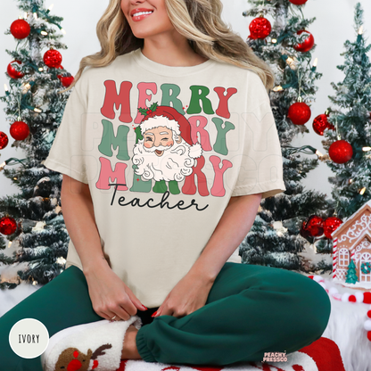 A woman wearing an ivory graphic t-shirt with a Christmas 'Merry Teacher' design, sitting in front of decorated Christmas trees. The t-shirt features a colorful retro-style graphic with Santa Claus and the text 'MERRY MERRY MERRY Teacher'.