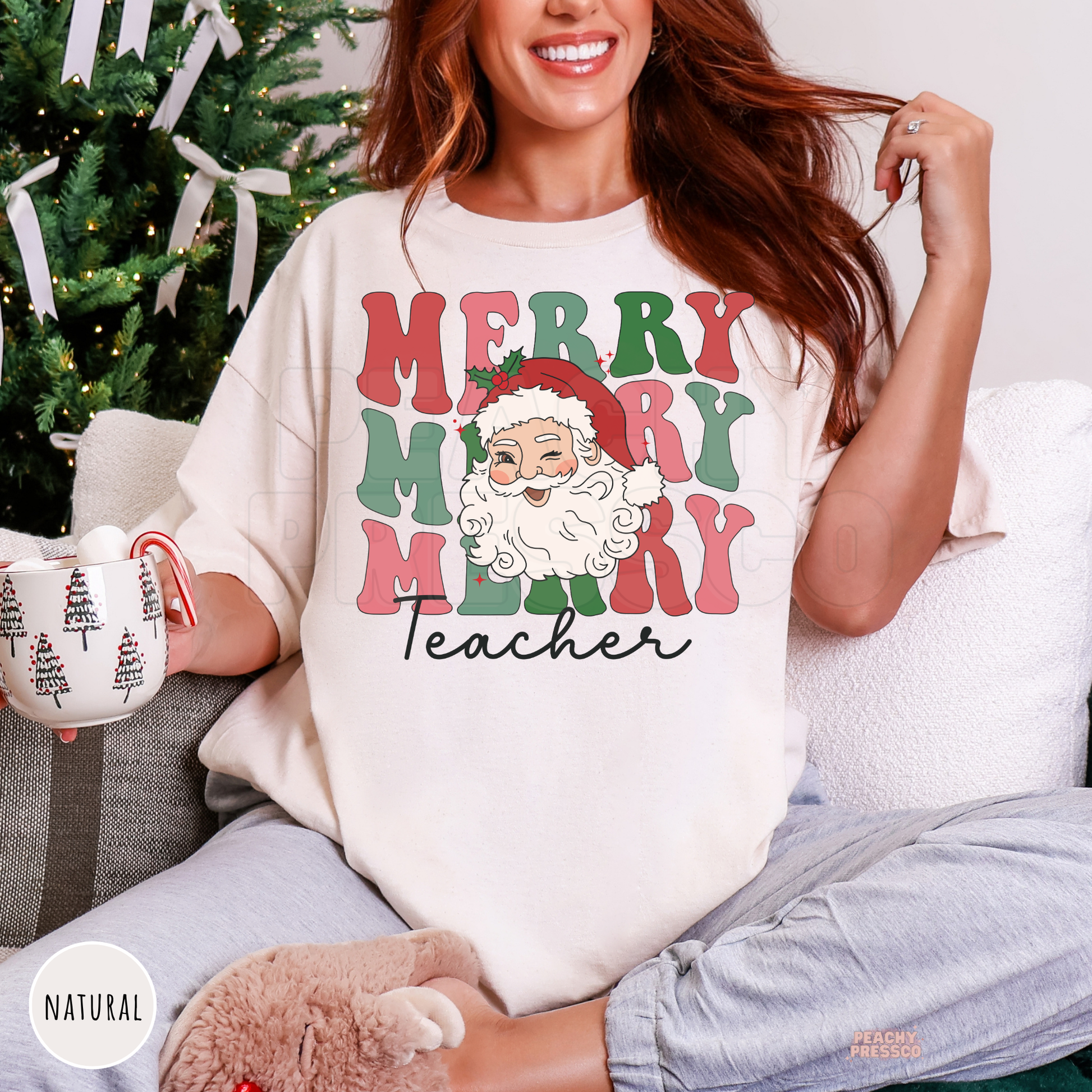 A woman sitting on a couch in front of a decorated Christmas tree wears an oversized natural colored graphic t-shirt that reads 'MERRY MERRY MERRY Teacher' with a Santa Claus illustration. She holds a mug and wears gray sweatpants.