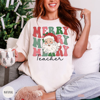A woman sitting on a couch in front of a decorated Christmas tree wears an oversized natural colored graphic t-shirt that reads 'MERRY MERRY MERRY Teacher' with a Santa Claus illustration. She holds a mug and wears gray sweatpants.