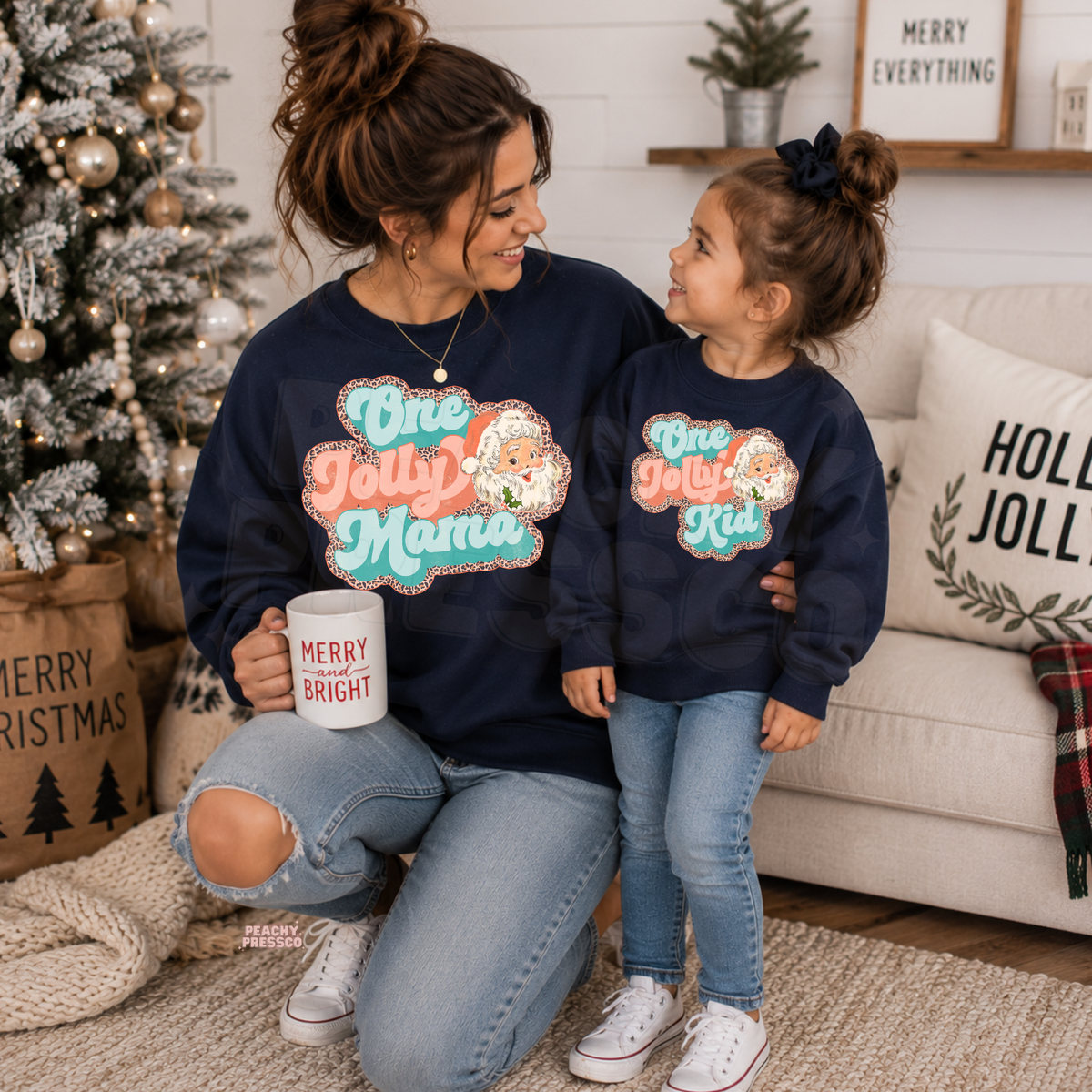 Mom and daughter wearing matching navy One Jolly Mama and One Jolly Kid Christmas crewneck sweatshirts by a Christmas tree.