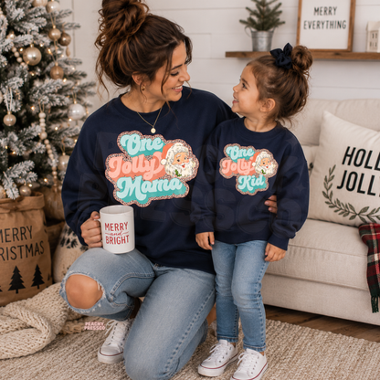 Mom and daughter wearing matching navy One Jolly Mama and One Jolly Kid Christmas crewneck sweatshirts by a Christmas tree.