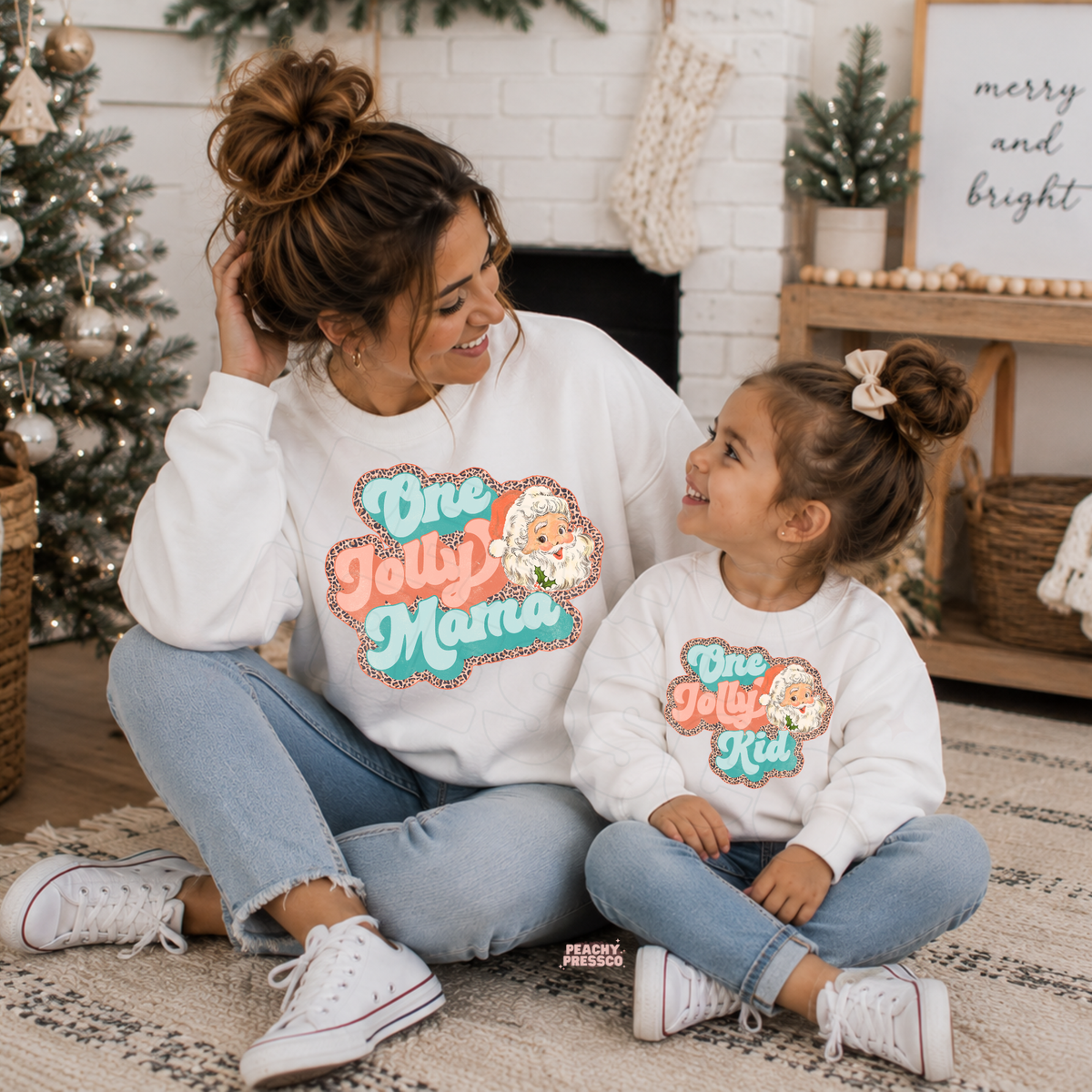 Mom and daughter wearing matching white One Jolly Mama and One Jolly Kid Christmas sweatshirts with retro Santa graphic.