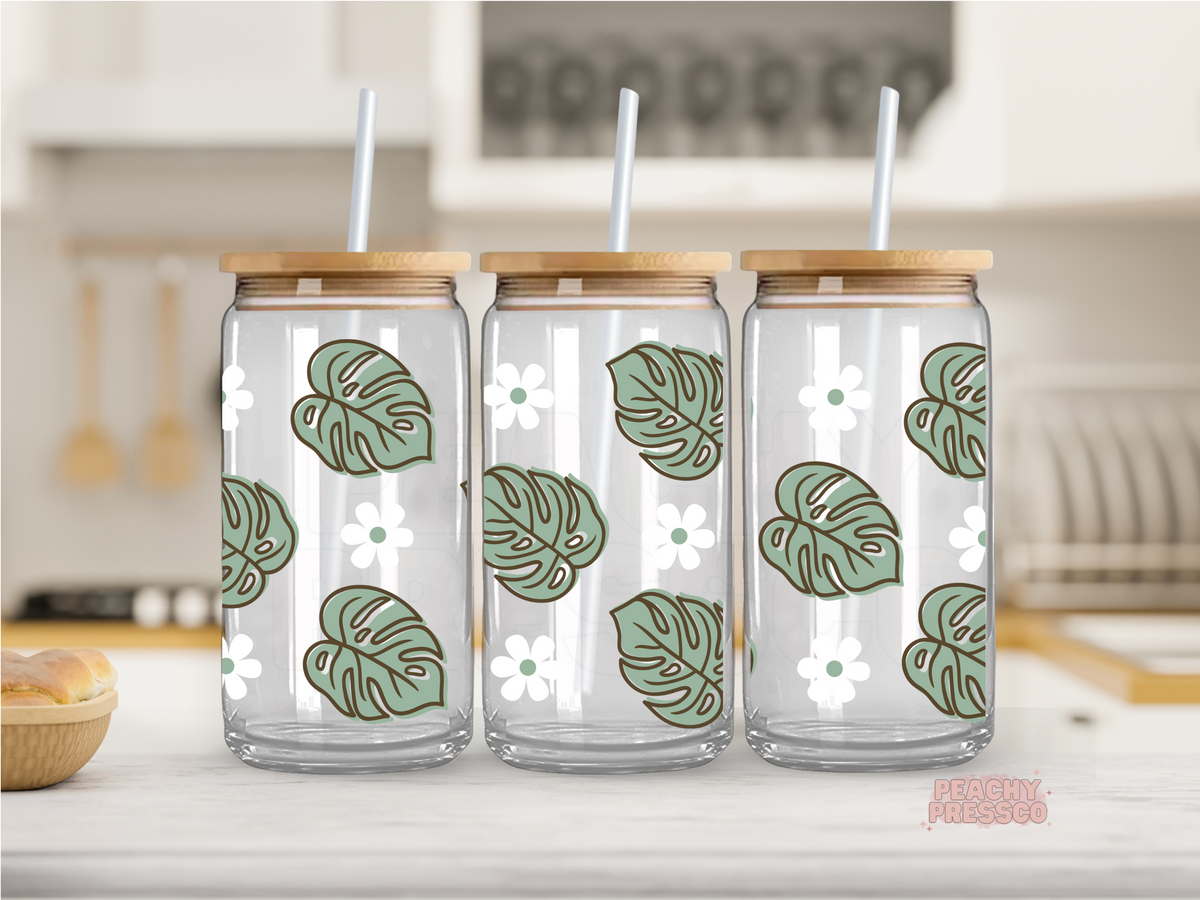 16 oz clear glass cup featuring sage green monstera leaves and white daisy flowers in a modern botanical pattern. Includes bamboo lid and reusable straw.