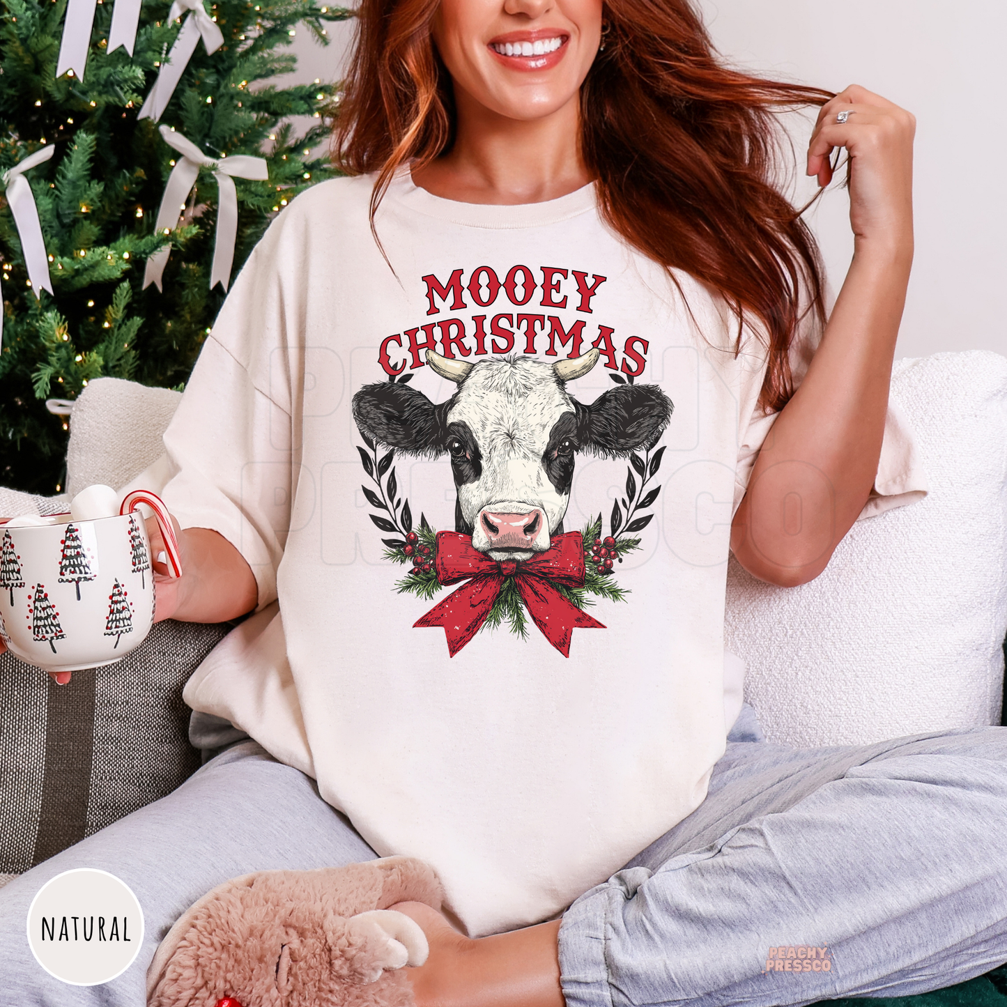 A woman wearing a natural colored Christmas graphic t-shirt with a cow and red bow design, sitting on a couch next to a decorated Christmas tree.