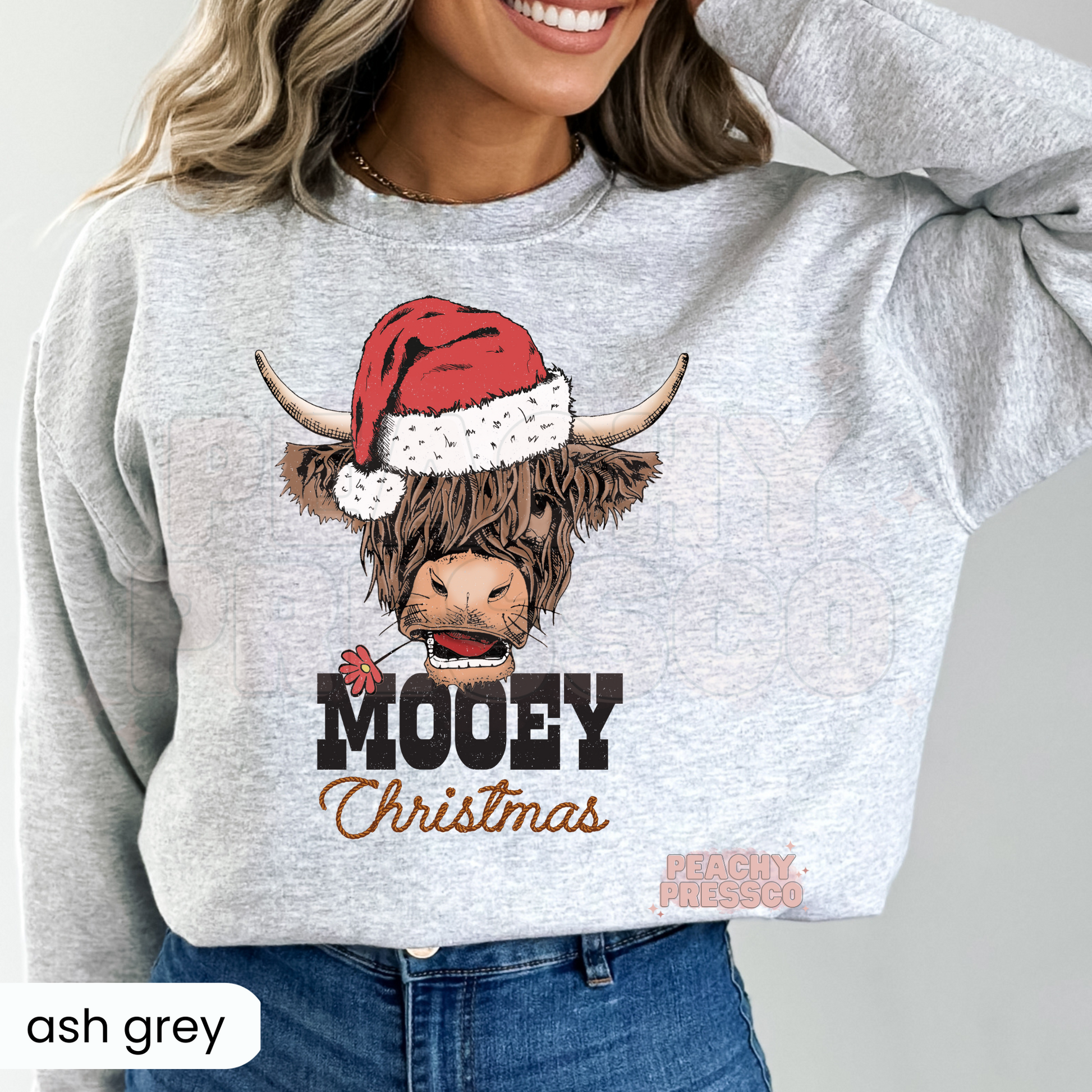 A woman wearing a ash gray sweatshirt with a graphic of a shaggy Highland cow in a Santa hat and the text 'MOOEY Christmas'.