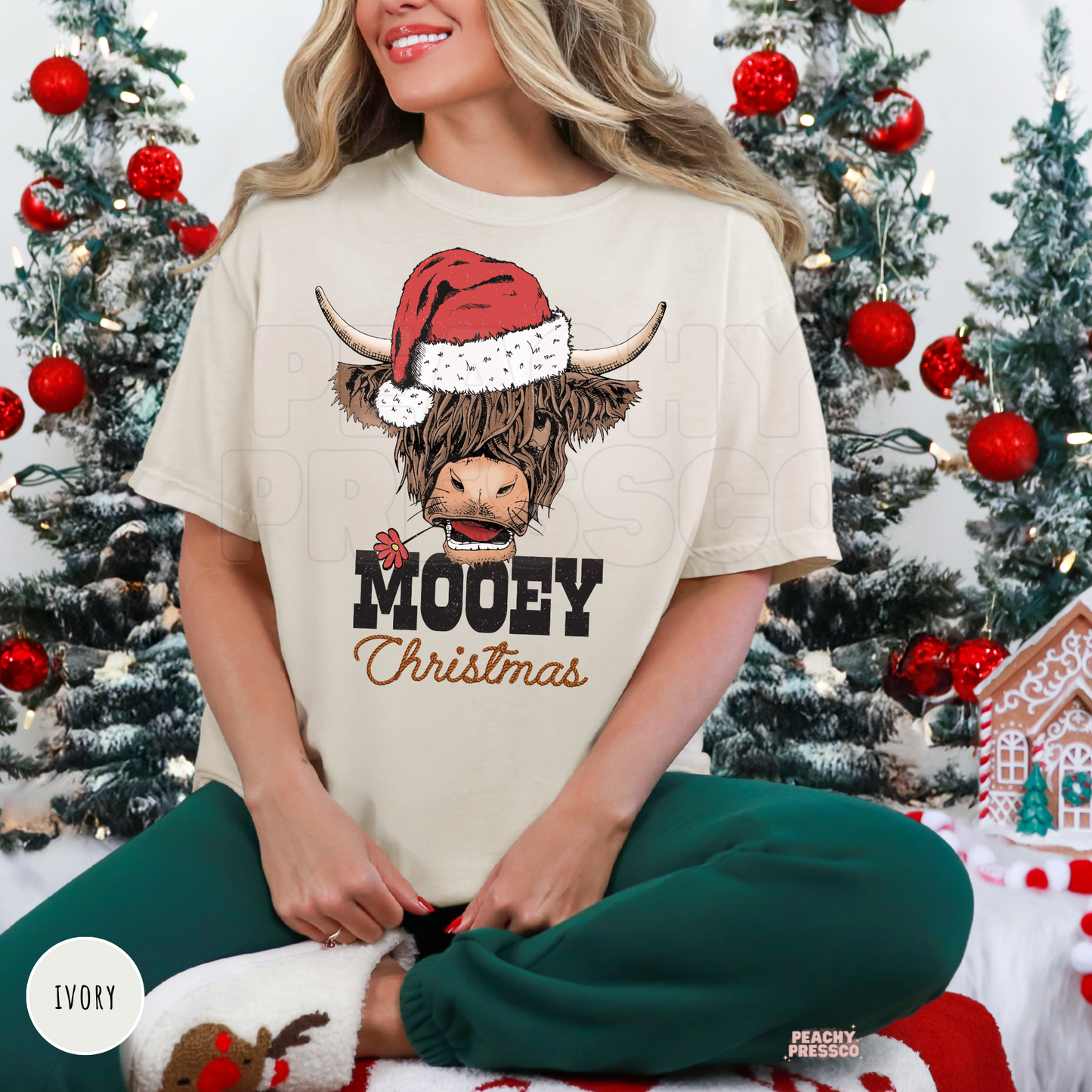 Woman sitting wearing an ivory Mooey Christmas highland cow t-shirt paired with dark green sweatpants.