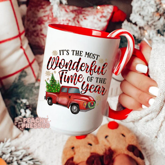 White holiday coffee mug with red handle featuring a red vintage pickup truck carrying a Christmas tree with plaid text reading Its The Most Wonderful Time Of The Year.