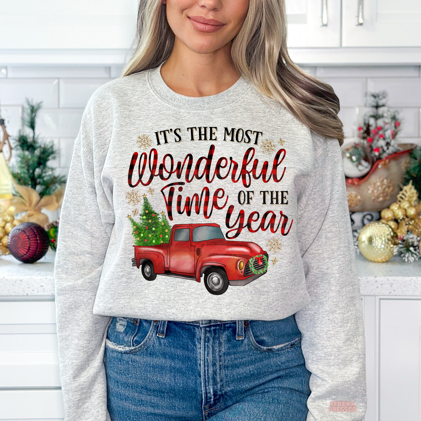 A woman wearing a ash grey crewneck sweatshirt with a festive graphic that reads 'IT'S THE MOST Wonderful Time OF THE YEAR' above a red vintage pickup truck carrying a Christmas tree, set against a decorated holiday kitchen background.