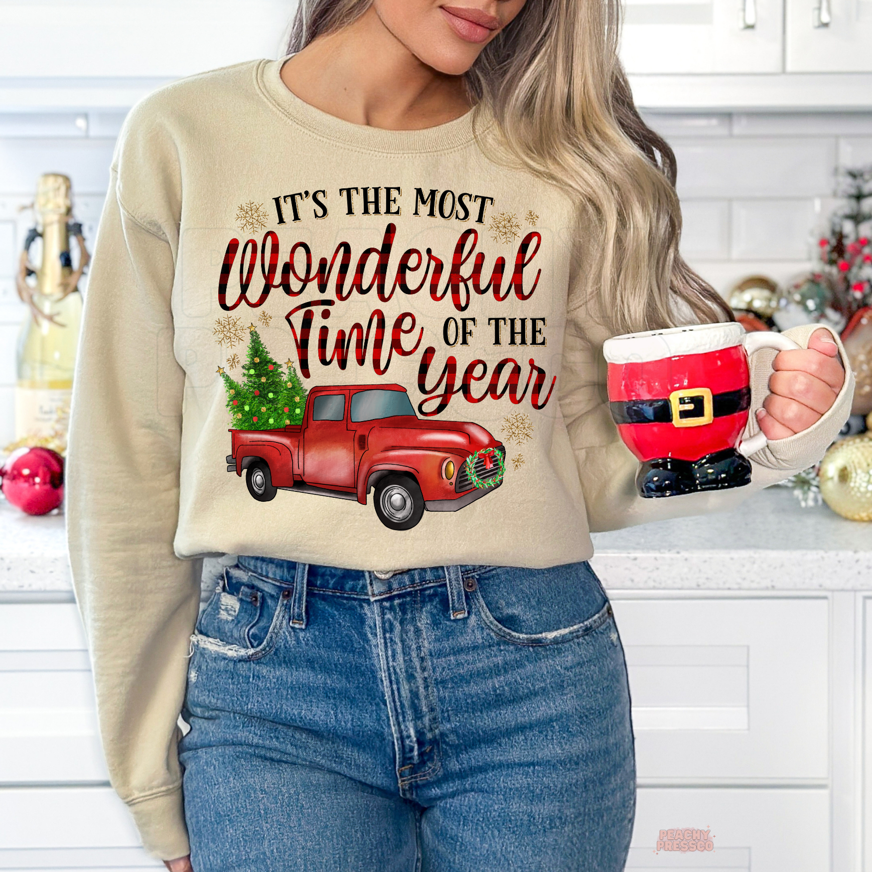 A woman wearing a sand crewneck sweatshirt with a Christmas graphic that reads 'IT'S THE MOST Wonderful Time OF THE year' above a red truck carrying a Christmas tree. She is holding a ceramic mug shaped like Santa Claus, set in a decorated holiday kitchen environment.