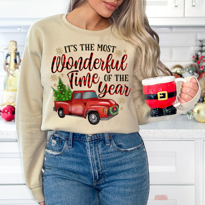 A woman wearing a sand crewneck sweatshirt with a Christmas graphic that reads 'IT'S THE MOST Wonderful Time OF THE year' above a red truck carrying a Christmas tree. She is holding a ceramic mug shaped like Santa Claus, set in a decorated holiday kitchen environment.