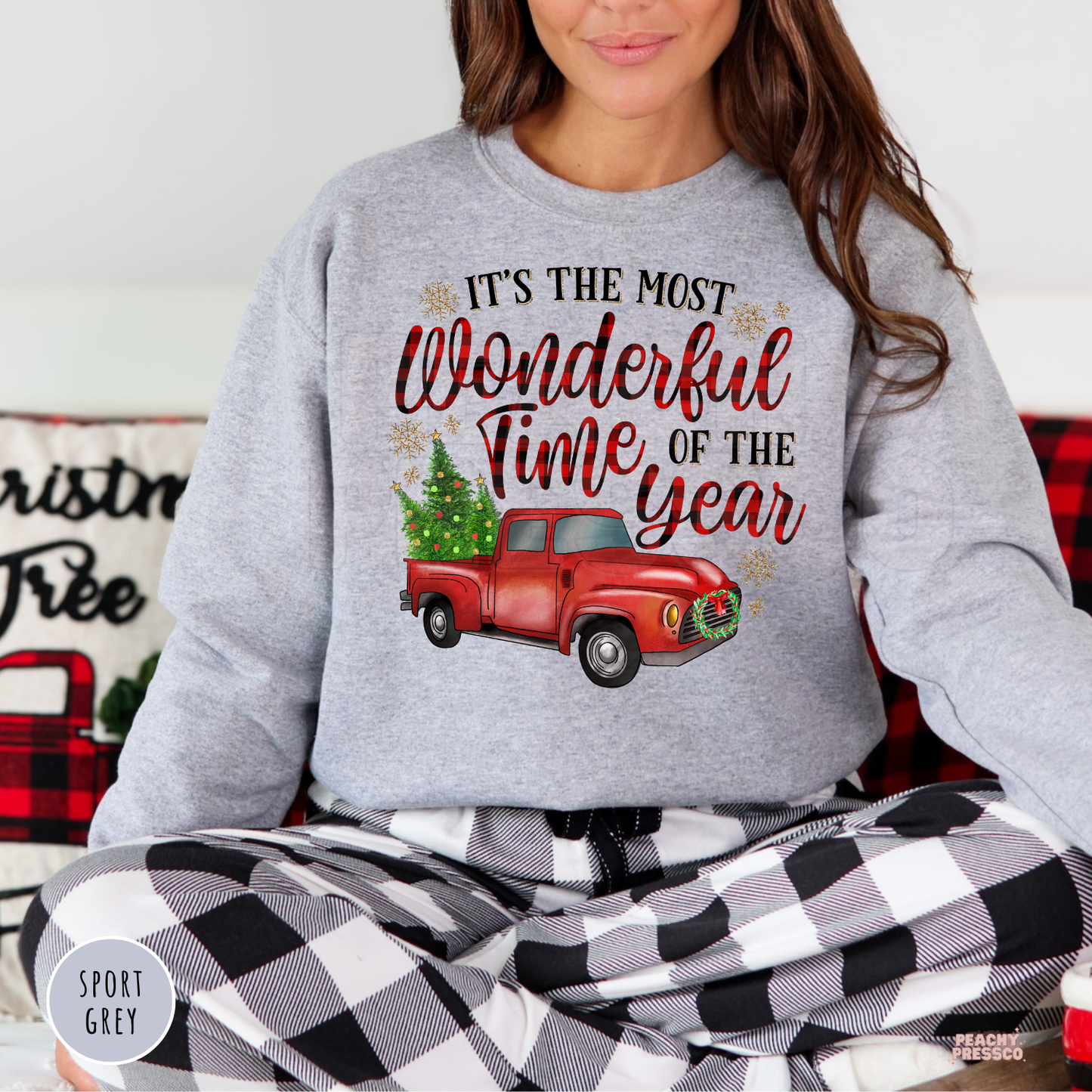 A woman wearing a sport grey sweatshirt with a red vintage truck graphic and the text 'It's the most wonderful time of the year,' paired with black-and-white checkered pajama pants, sitting indoors.