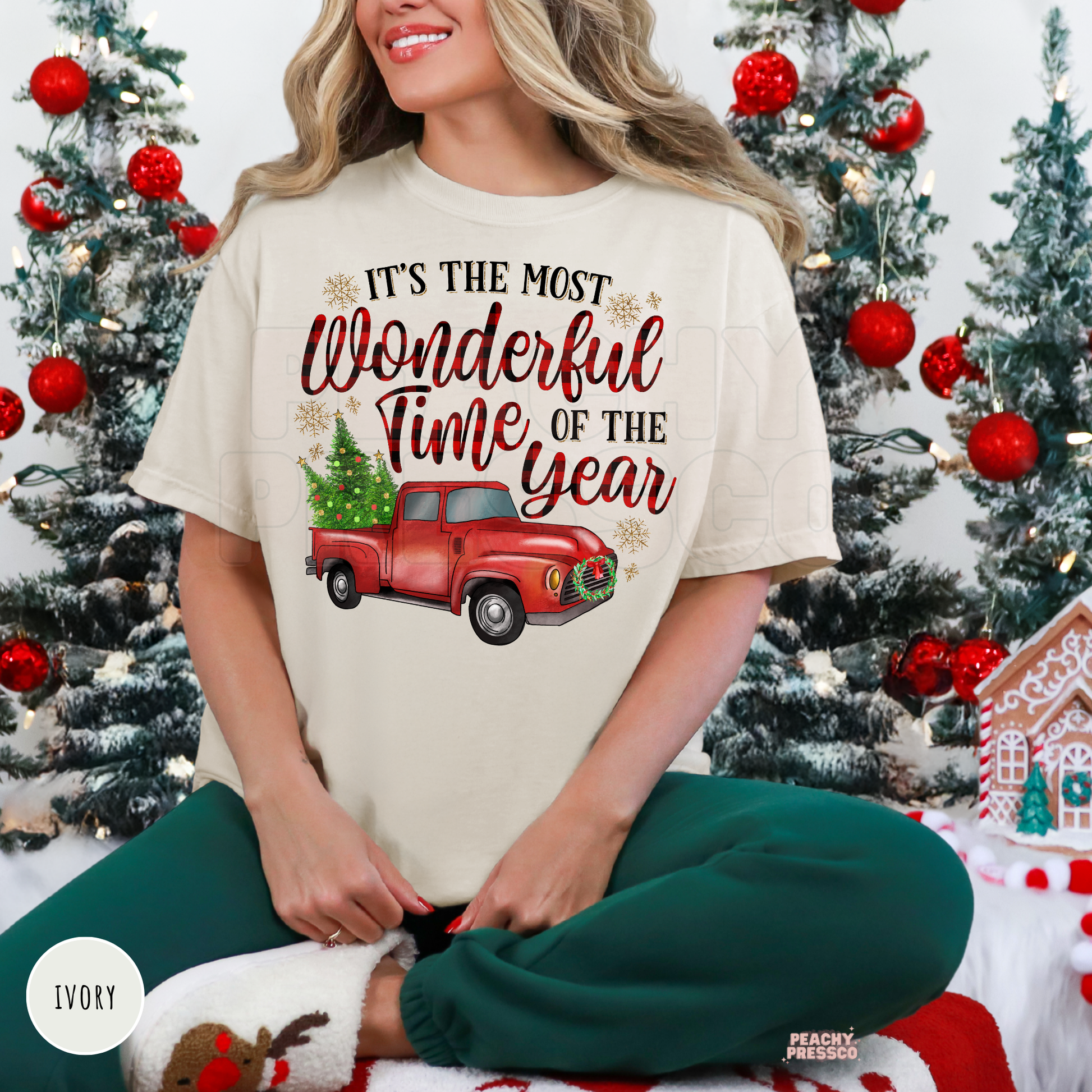 A woman wearing an ivory graphic t-shirt with a red truck and Christmas tree design, paired with green pants, sitting in front of decorated Christmas trees. The t-shirt features the text 'IT'S THE MOST Wonderful Time OF THE year'.
