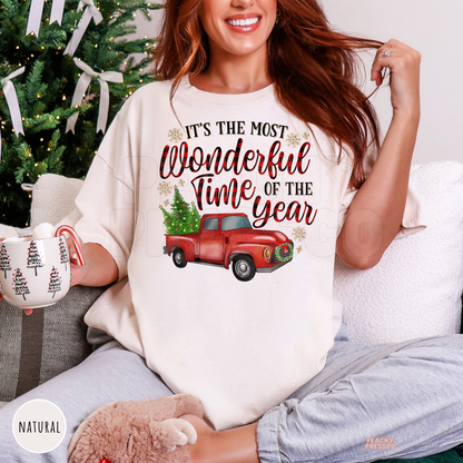 A woman sitting on a couch wearing a natural colored graphic t-shirt with a red vintage truck carrying a Christmas tree and the text 'IT'S THE MOST Wonderful Time OF THE YEAR'. She is holding a mug in front of a decorated Christmas tree.