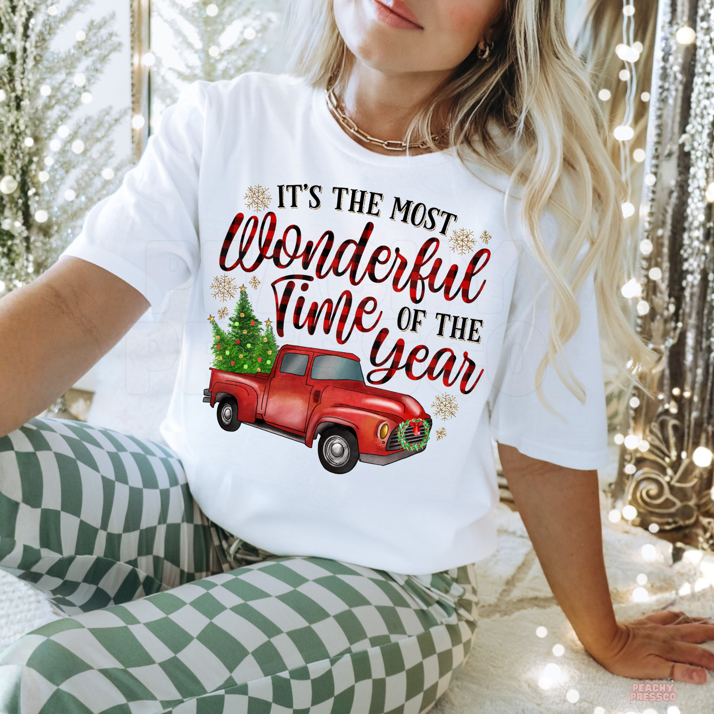 A woman wearing a white graphic t-shirt with a red vintage truck carrying a Christmas tree and the text 'IT'S THE MOST Wonderful Time OF THE YEAR', paired with green and white checkered pajama pants in a cozy holiday setting with string lights.