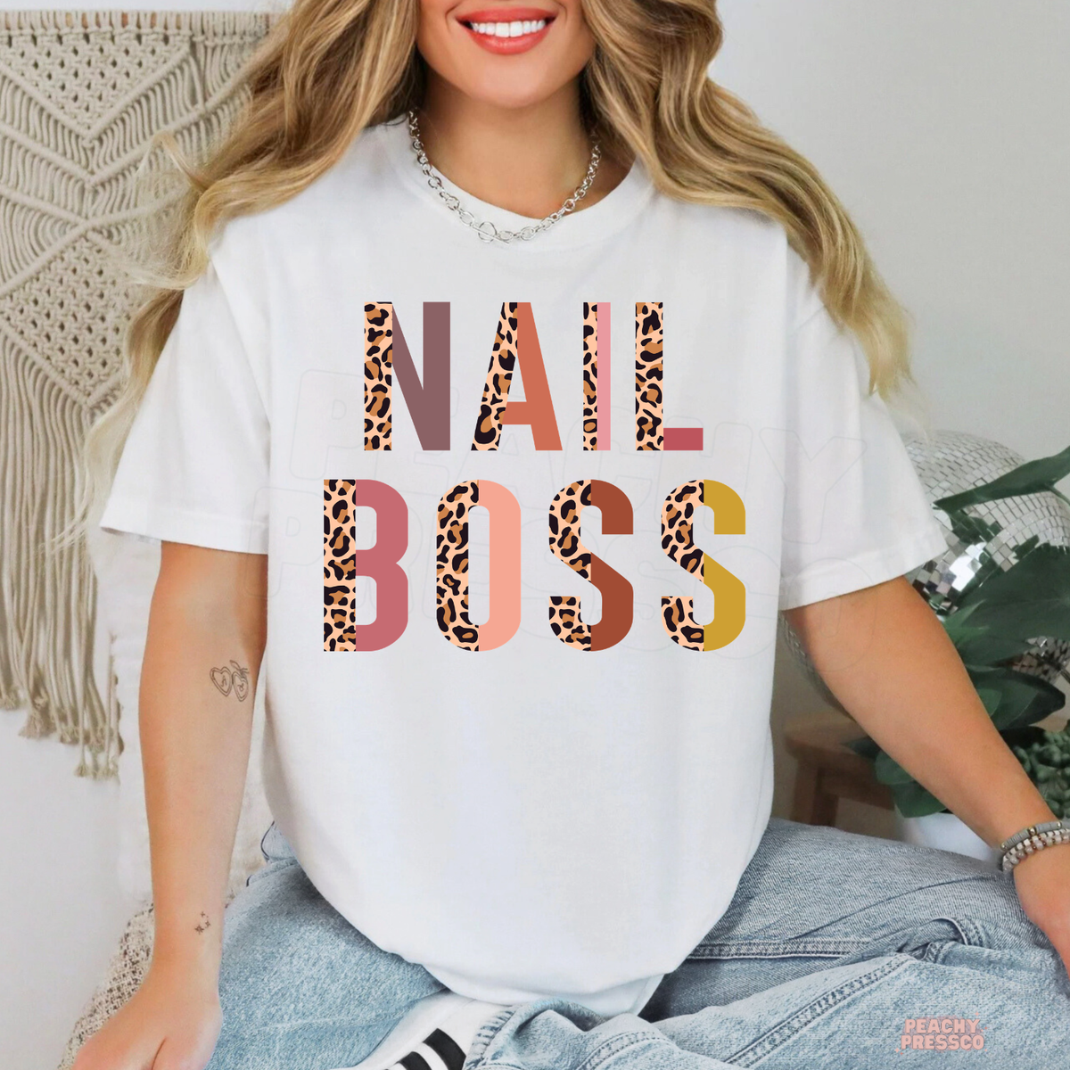 Woman modeling a white Nail Boss T-shirt featuring bold leopard print letters in pink, coral, brown and mustard tones.