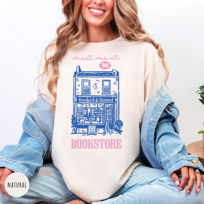 Natural cream tee featuring a charming bookstore illustration with pink and blue "Meet Me at the Bookstore" graphic. Perfect cozy shirt for readers and book lovers.