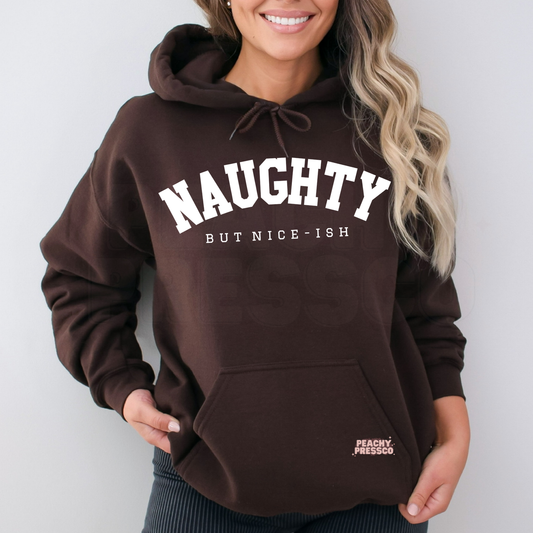 A woman wearing a dark brown pullover hoodie with 'NAUGHTY BUT NICE-ISH' printed in white on the front. The hoodie has a drawstring hood and a pocket.