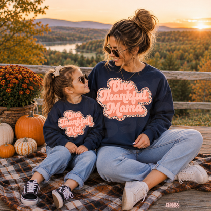 Mother and daughter in matching navy One Thankful Mama and One Thankful Kid sweatshirts overlooking colorful autumn scenery at sunset.