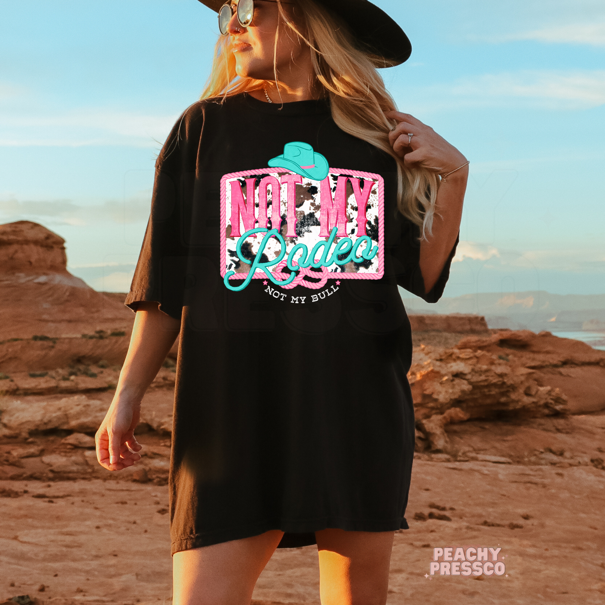Woman wearing a black t-shirt featuring a funny "Not My Rodeo, Not My Bull" western graphic with turquoise cowboy hat and cow print design.