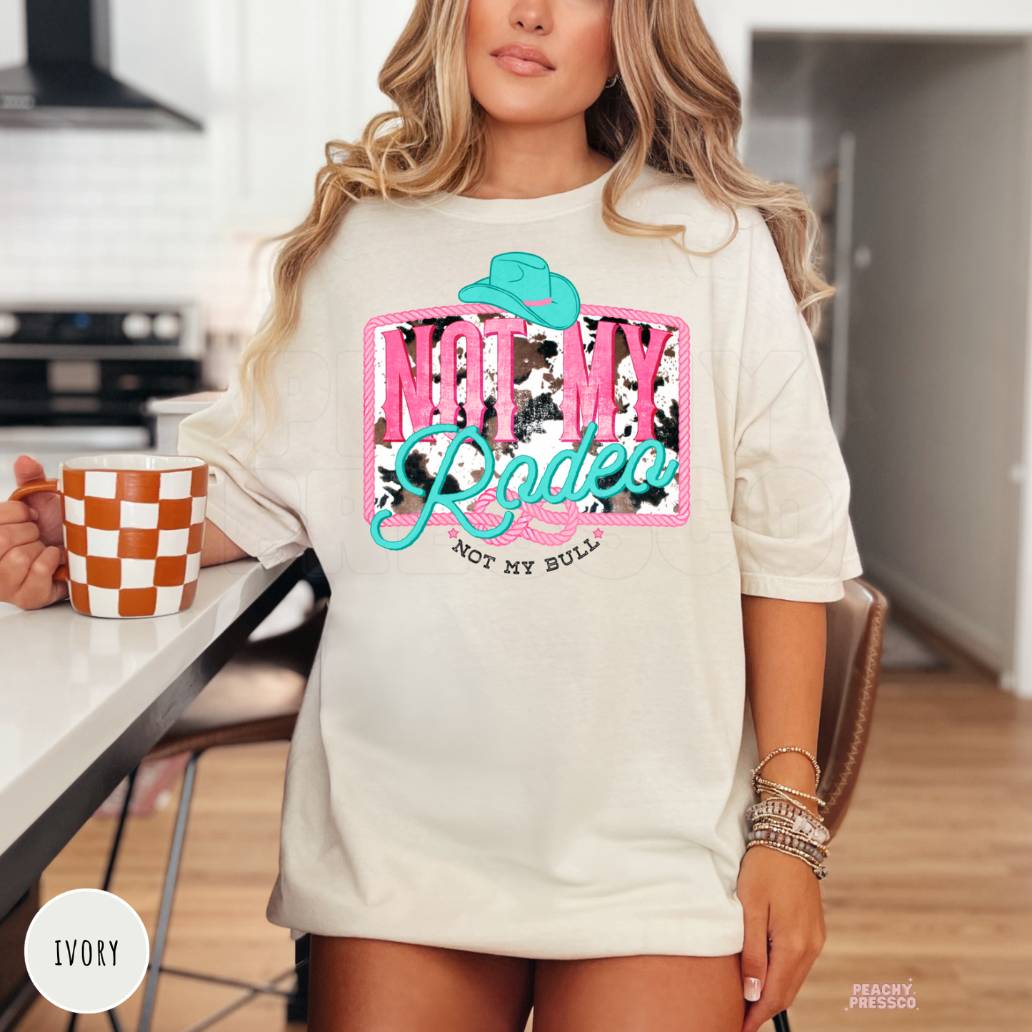 Woman wearing an ivory t-shirt with a vibrant "Not My Rodeo, Not My Bull" western cowgirl graphic featuring cow print, turquoise cowboy hat, and pink lettering.