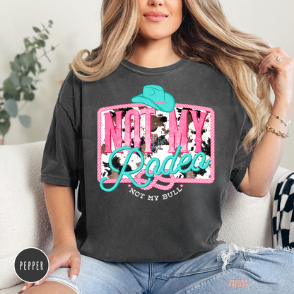Woman wearing a pepper Comfort Colors t-shirt featuring a funny "Not My Rodeo, Not My Bull" western graphic with turquoise cowboy hat and pink cowgirl design.