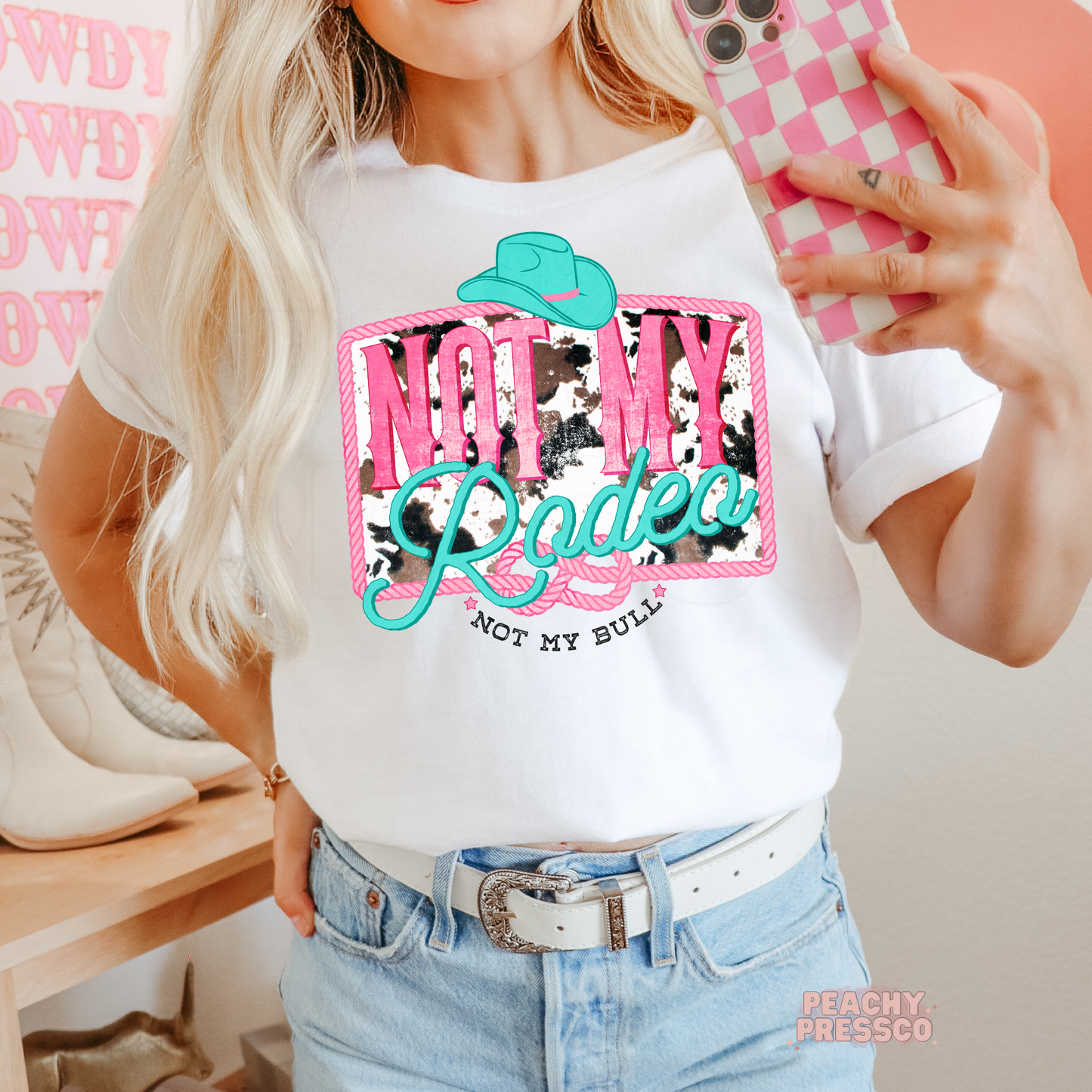Woman wearing a white t-shirt featuring a bold "Not My Rodeo, Not My Bull" western cowgirl graphic with turquoise cowboy hat and pink lettering.